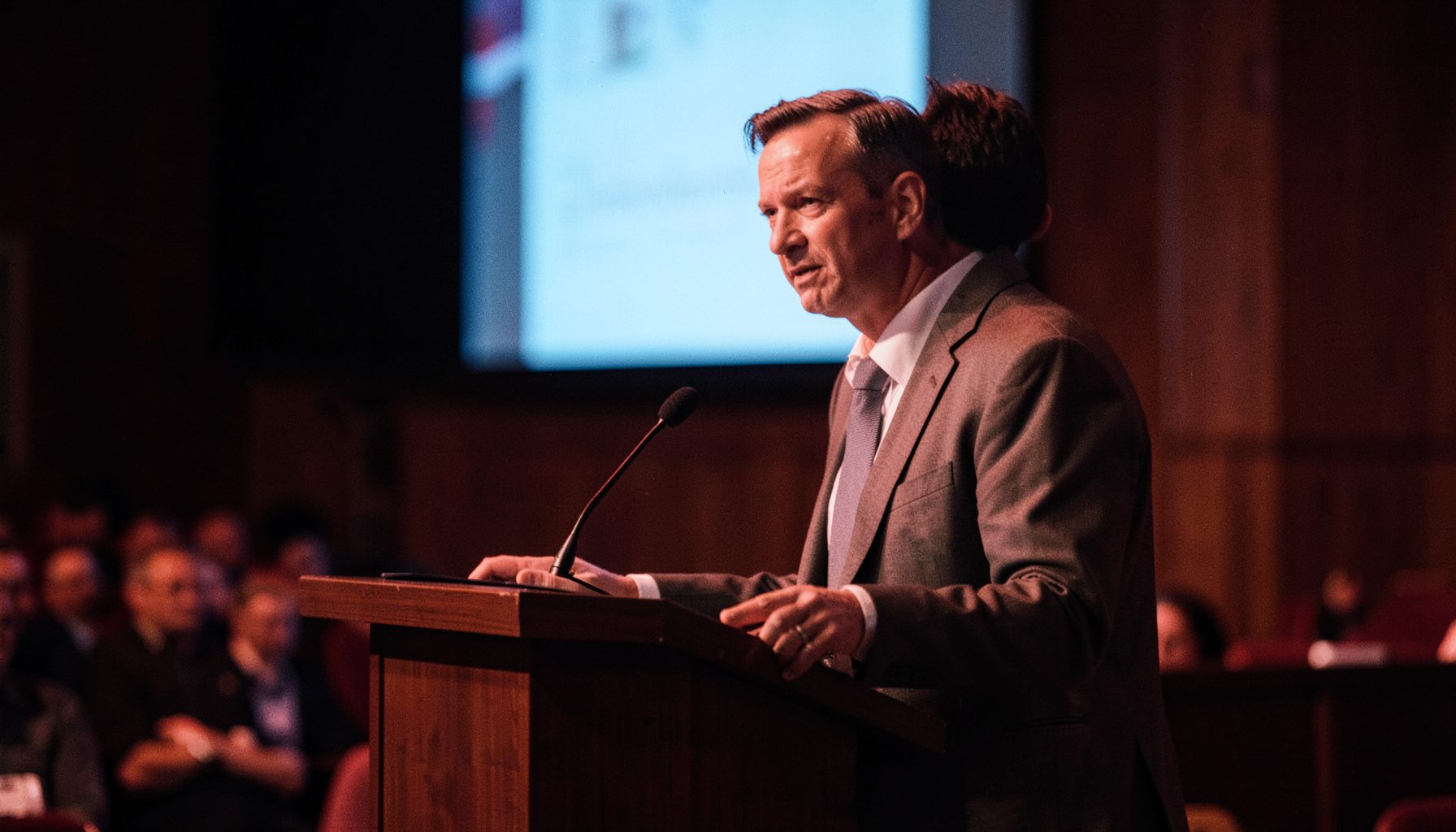 A health-sector keynote speaker at a podium in a softly lit auditorium, wearing professional attire, with a presentation screen glowing behind them
