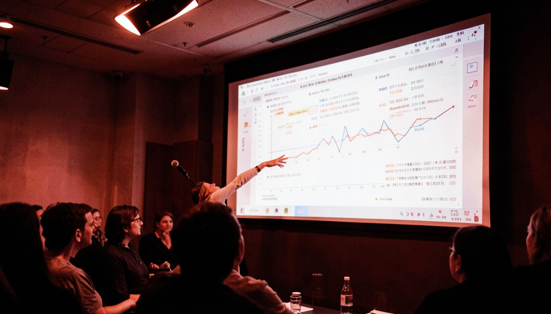 A science-sector presenter gesturing toward a data-rich slide on a large screen, with warm overhead lighting and a focused audience