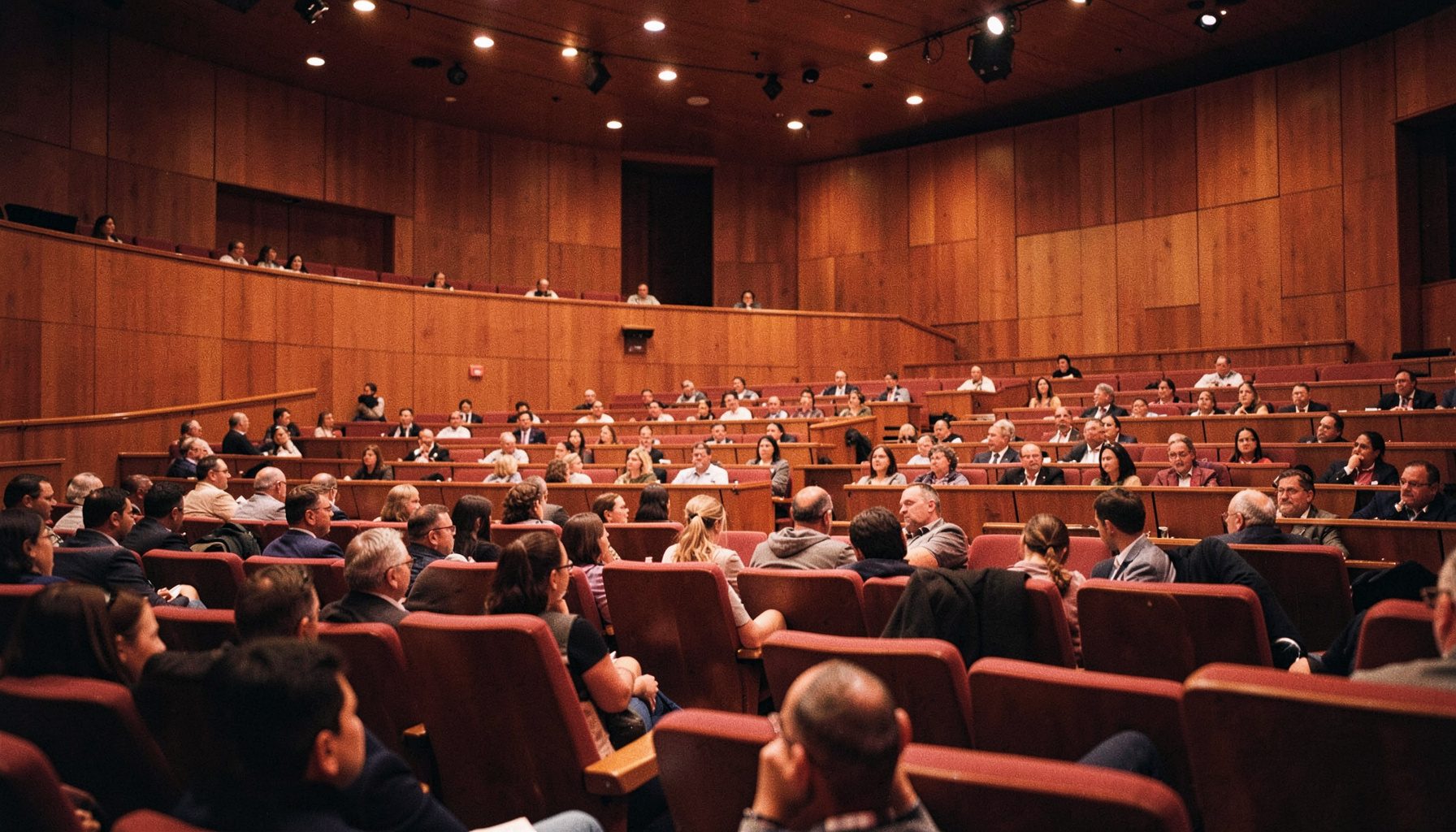A wide conference lecture hall with warm wood paneling, rows of tiered seating, and soft amber stage lighting casting a golden glow