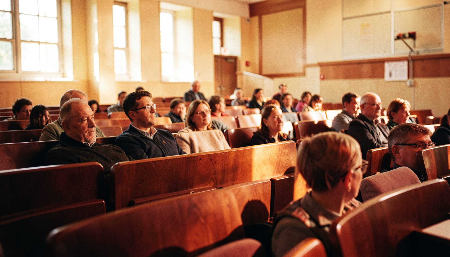 A sunlit university lecture hall with rows of wooden seats, warm amber and cream tones, a sense of quiet anticipation before a conference session