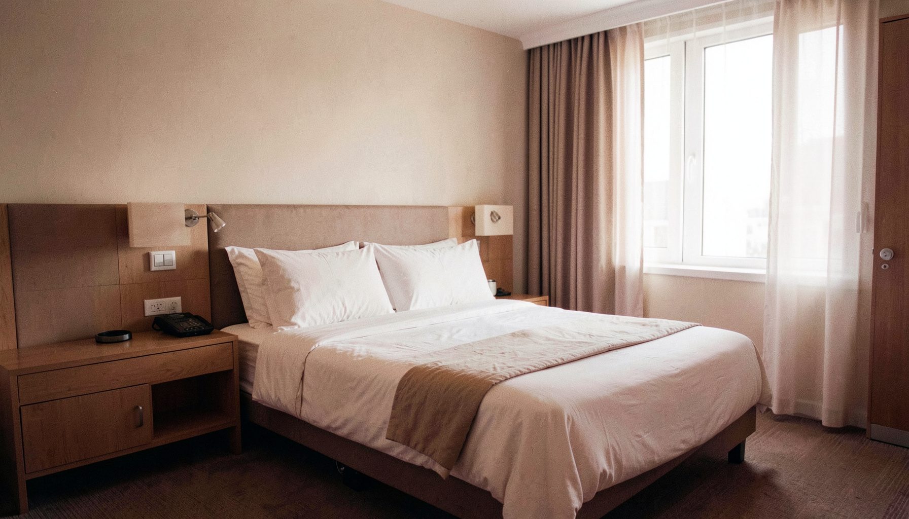 A bright modern hotel room with a neatly made bed, warm neutral tones, and soft daylight through a window